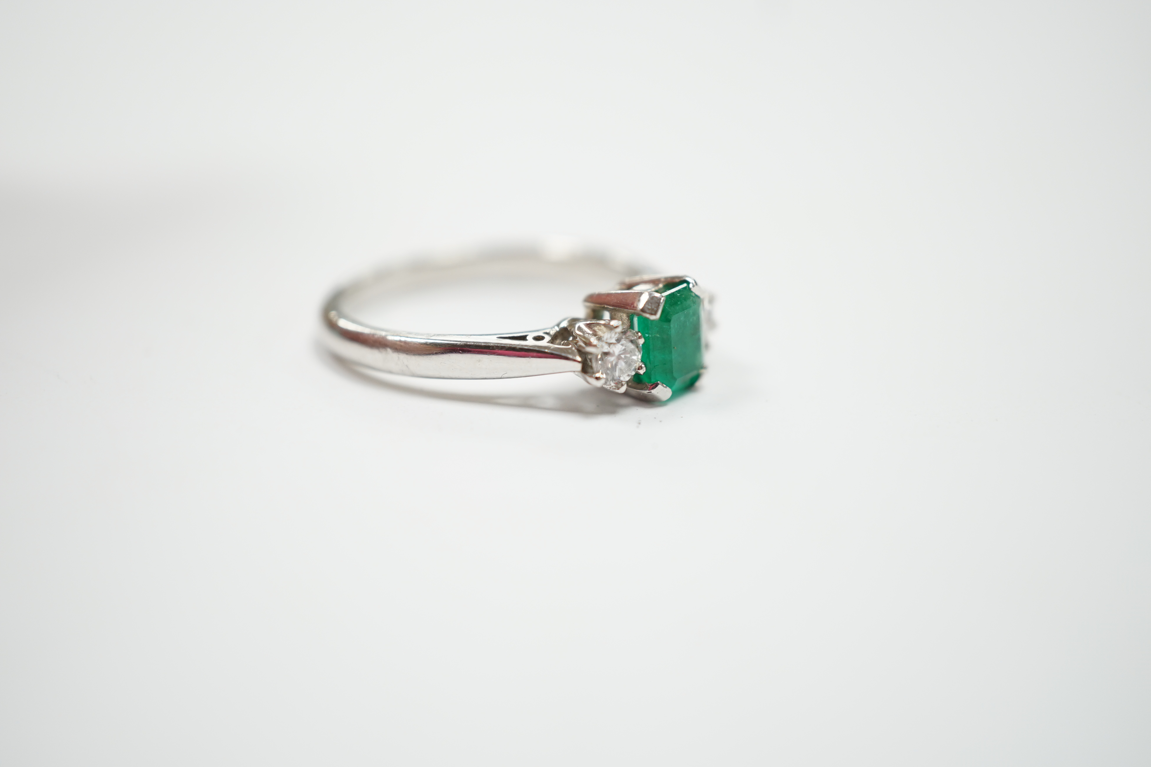 A modern 18ct white metal, single stone emerald and two stone diamond set ring, size N, gross weight 4 gram.